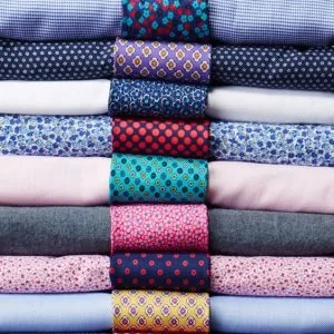 Stack of premium custom dress shirts in various colors with matching patterned silk ties, representing exclusive fabric choices and bespoke accessories at From Collars to Cuffs in South Florida
