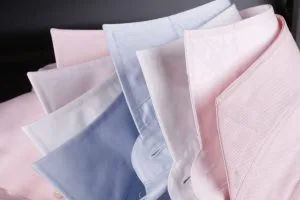 Stack of neatly folded custom dress shirts in pastel pink, light blue, white, and lavender, showcasing premium cotton fabric quality and color variety for bespoke menswear at From Collars to Cuffs in South Florida