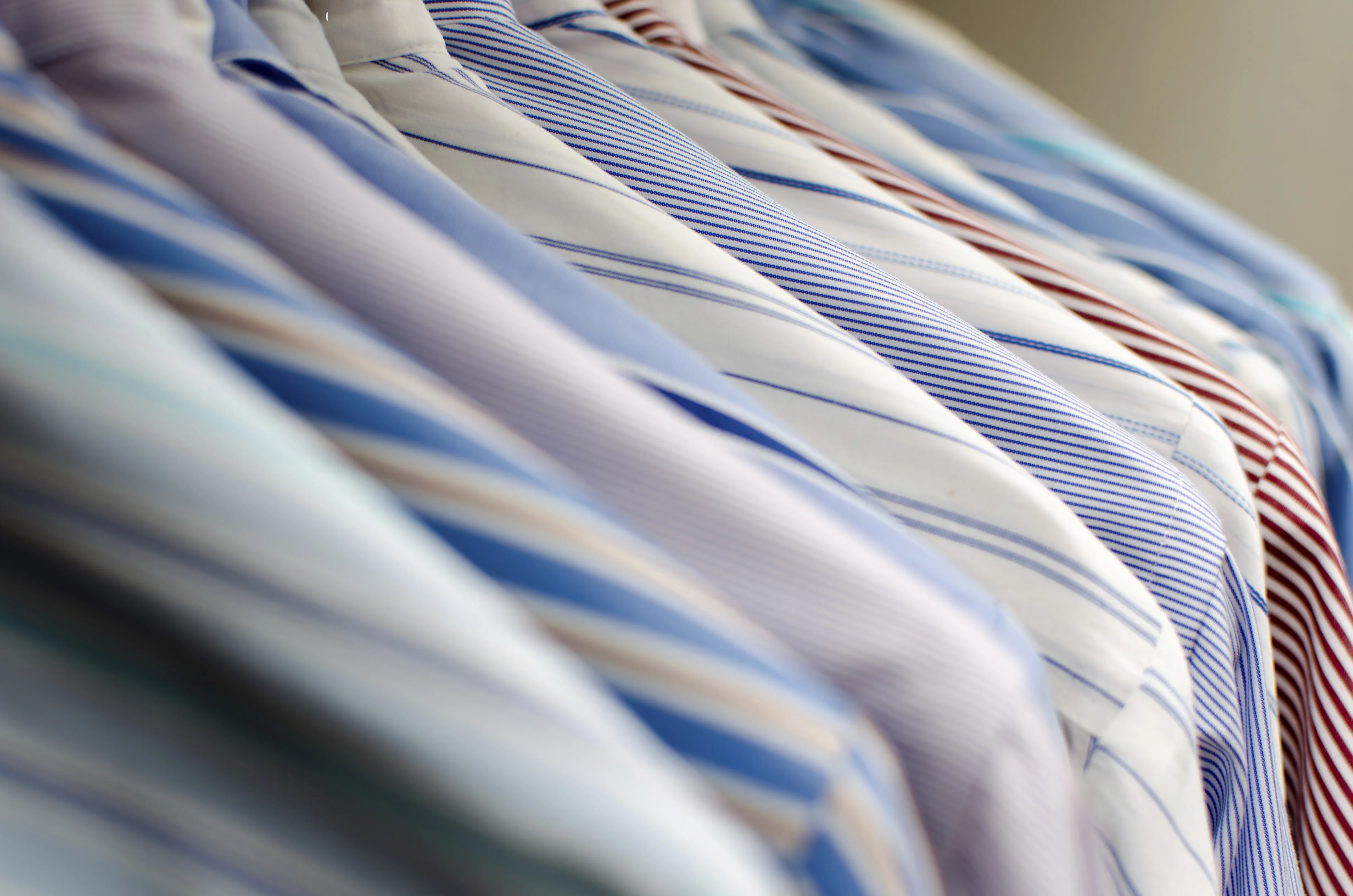 Row of hanging custom dress shirts in various striped and solid patterns, highlighting the extensive fabric library and personalized shirt options available for bespoke orders at From Collars to Cuffs in Weston, Florida