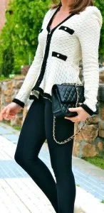 Woman modeling a custom white textured jacket with black trim and buttons, paired with black pants and a quilted handbag, representing bespoke Chanel-inspired fashion at From Collars to Cuffs.