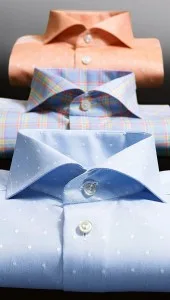 Collection of custom-tailored men's dress shirts in peach, blue plaid, and light blue, showcasing a variety of premium fabric colors and patterns available for bespoke orders at From Collars to Cuffs in Florida