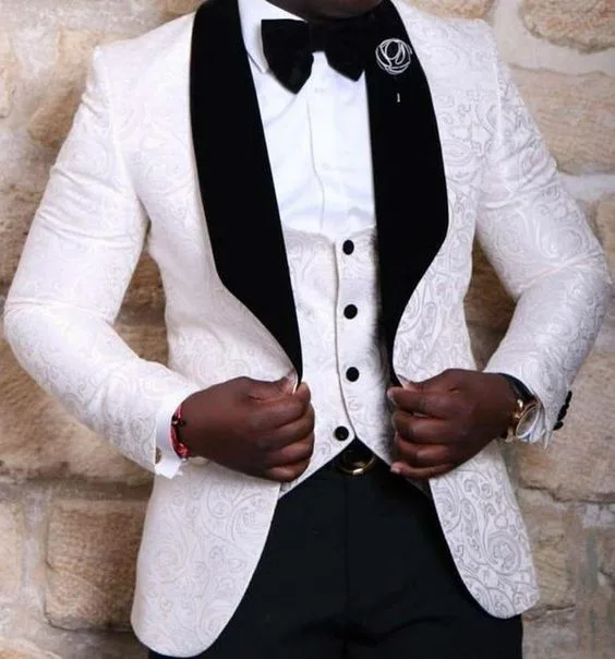 Man wearing a custom white textured tuxedo jacket with black velvet lapels and bow tie against a stone wall, representing elegant bespoke formal wear at From Collars to Cuffs in South Florida.