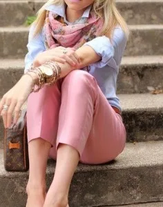 Woman sitting on steps wearing custom pink trousers, a light blue shirt, and a silk scarf, demonstrating bespoke styling and casual elegance available at From Collars to Cuffs.