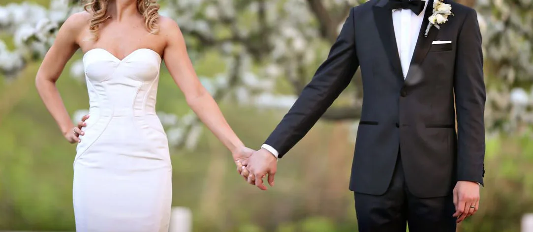 Bride in custom white wedding dress and groom in bespoke black tuxedo holding hands, showcasing luxury wedding attire tailoring at From Collars to Cuffs in South Florida