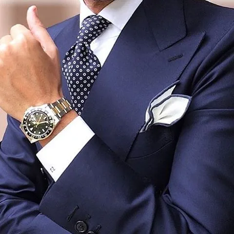 Close-up of a man in a navy blue suit adjusting his tie and cuff, highlighting premium fabric texture, cufflinks, and luxury watch accessories for custom menswear at From Collars to Cuffs in Florida
