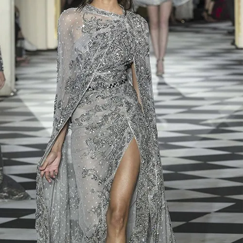 Model walking in a custom silver sheer cape gown with beaded embellishments and a high leg slit, highlighting elegant bespoke formal wear for galas at From Collars to Cuffs in South Florida