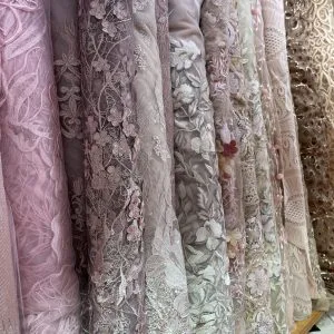 Close-up of elegant lace and embroidered fabric rolls in soft pinks and whites, showcasing premium textile choices for custom women's dresses, blouses, and gowns at From Collars to Cuffs.