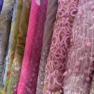 Display of premium fabric bolts featuring pink lace, green embroidered details, and sheer materials with 3D floral appliqués, showcasing luxury textile options for custom women's fashion at From Collars to Cuffs in Weston, South Florida.
