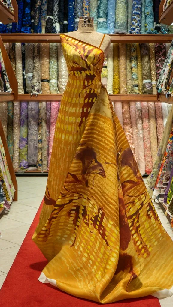 Mannequin wearing a custom gold and yellow silk print cocktail dress surrounded by luxury fabric rolls, highlighting bespoke women's fashion and exclusive material options at From Collars to Cuffs in Weston, Florida