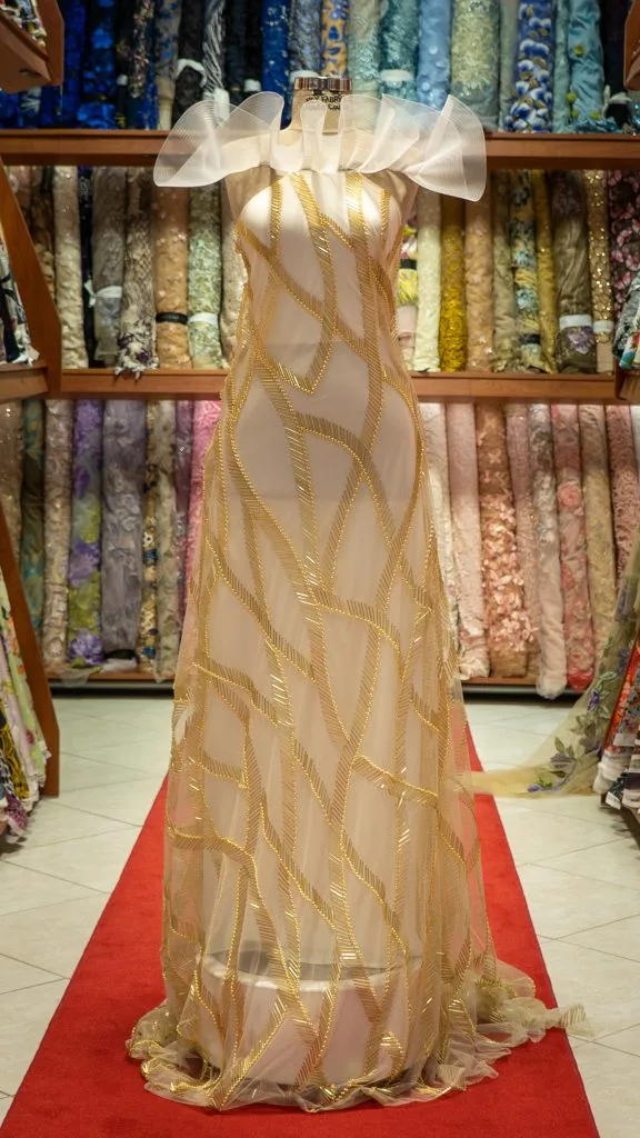 Mannequin displaying a custom sheer evening gown with gold embroidery in a boutique fabric showroom, showcasing bespoke women's black tie gowns and premium textile selection at From Collars to Cuffs in South Florida