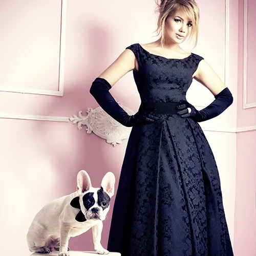 Sophisticated woman wearing elegant black brocade evening gown with long black gloves, standing with French bulldog at From Collars to Cuffs luxury fashion boutique in Weston, Florida
